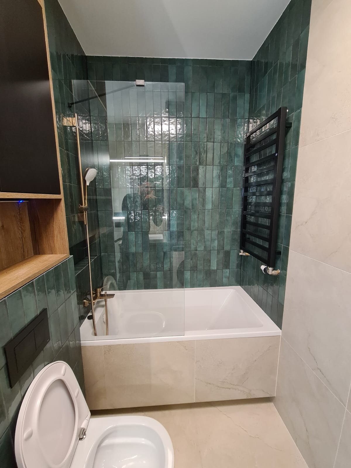 Nowoczesna łazienka z wanną obudowaną beżowymi płytami imitującymi marmur, zielone pionowe płytki na ścianie, czarny grzejnik drabinkowy i armatura w kolorze starego złota.