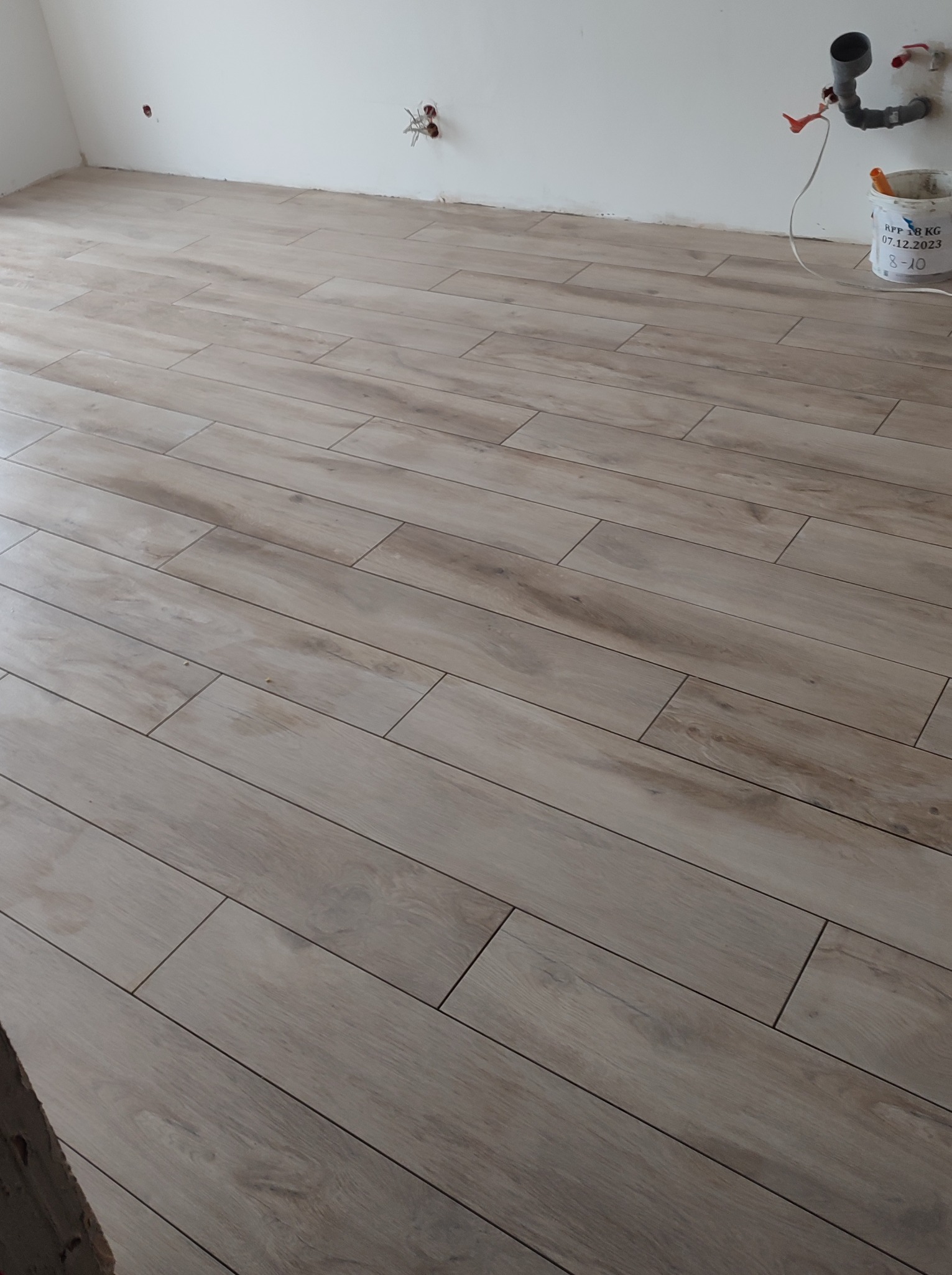 Ułożone pod kątem płytki podłogowe imitujące drewno, z widocznymi fugami, na tle białej ściany z wystającymi kablami i rurami oraz wiadrem z farbą.