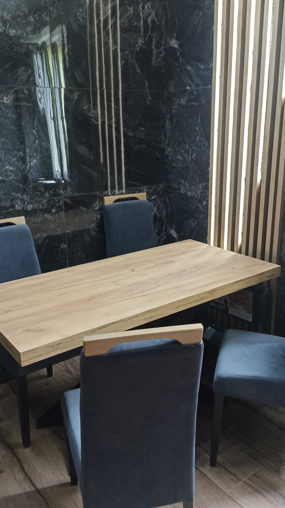 Drewniany stół z krzesłami z ciemnoszarą tapicerką na tle ściany z czarnych marmurowych płytek i drewnianych paneli lamelowych.