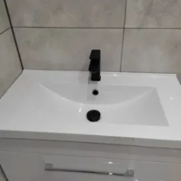 Biała umywalka z szafką i czarną, minimalistyczną armaturą na tle szarych, ceramicznych płytek imitujących beton.