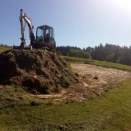 Mała koparka na wzniesieniu z ziemi, przygotowany teren pod budowę widoczny w tle, drzewa w oddali, słoneczny dzień.