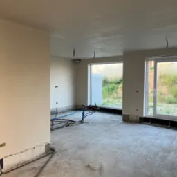 Wnętrze w stanie surowym z częściowo nałożonymi tynkami na ścianach, widoczne instalacje elektryczne i hydrauliczne, okna z widokiem na zieleń na zewnątrz.