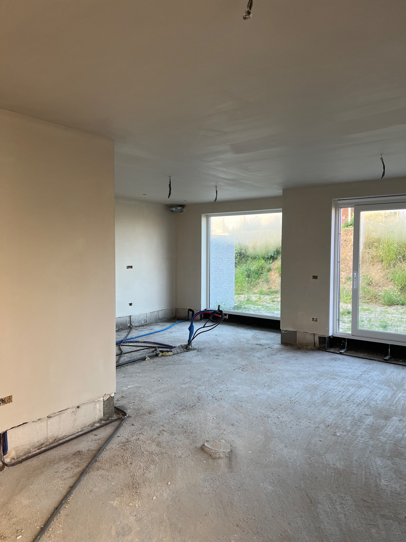 Wnętrze w stanie surowym z częściowo nałożonymi tynkami na ścianach, widoczne instalacje elektryczne i hydrauliczne, okna z widokiem na zieleń na zewnątrz.