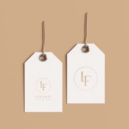 MIXTURE MARKETING SPÓŁKA Z OGRANICZONĄ ODPOWIEDZIALNOŚCIĄ - Dwie białe etykiety z logo 'LF' w okręgu, zawieszone na sznurkach na jednolitym, beżowym tle.