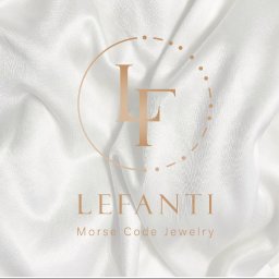 MIXTURE MARKETING SPÓŁKA Z OGRANICZONĄ ODPOWIEDZIALNOŚCIĄ - Logo firmy Lefanti Morse Code Jewelry na tle białej, jedwabnej tkaniny z widocznymi fałdami.