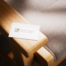 MIXTURE MARKETING SPÓŁKA Z OGRANICZONĄ ODPOWIEDZIALNOŚCIĄ - Wizytówka Glamour Academy z logo i tekstem 'Szkolenia z fryzur i makijażu' leży na drewnianym podłokietniku fotela obitego szarą tkaniną.