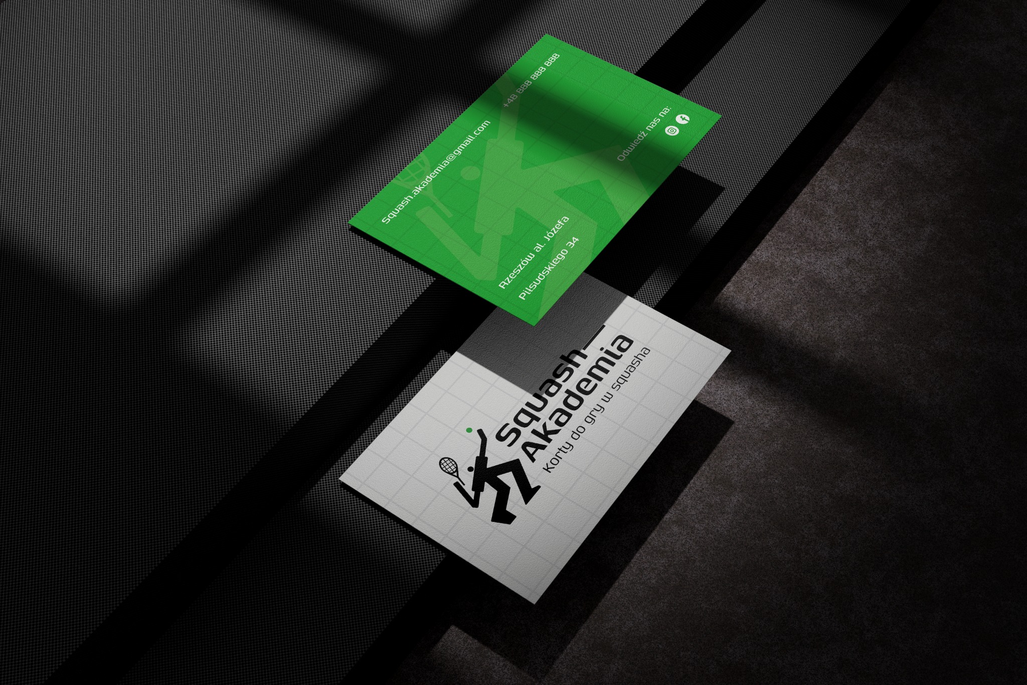 Dwie wizytówki Squash Akademii na ciemnym tle z cieniem: zielona z danymi kontaktowymi i biała z logo oraz informacją o kortach do gry w squasha.