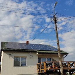 Artur Kutra Eko Monters - Biały dom z panelem słonecznym na dachu i drewnianym balkonem, widok z dołu na tle błękitnego nieba z chmurami, z przodu ogrodzenie z cegły i żelaza.