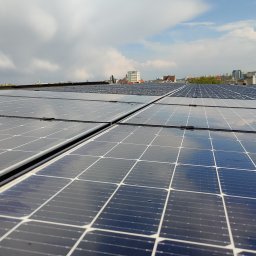 Artur Kutra Eko Monters - Panel słoneczny na dachu budynku, z widocznym odbiciem chmur i fragmentem panoramy miasta w tle.