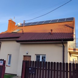 Artur Kutra Eko Monters - Montaż paneli słonecznych na dachu domu jednorodzinnego z czerwoną dachówką, widoczny pracownik w białej koszulce na dachu, błękitne niebo w tle.