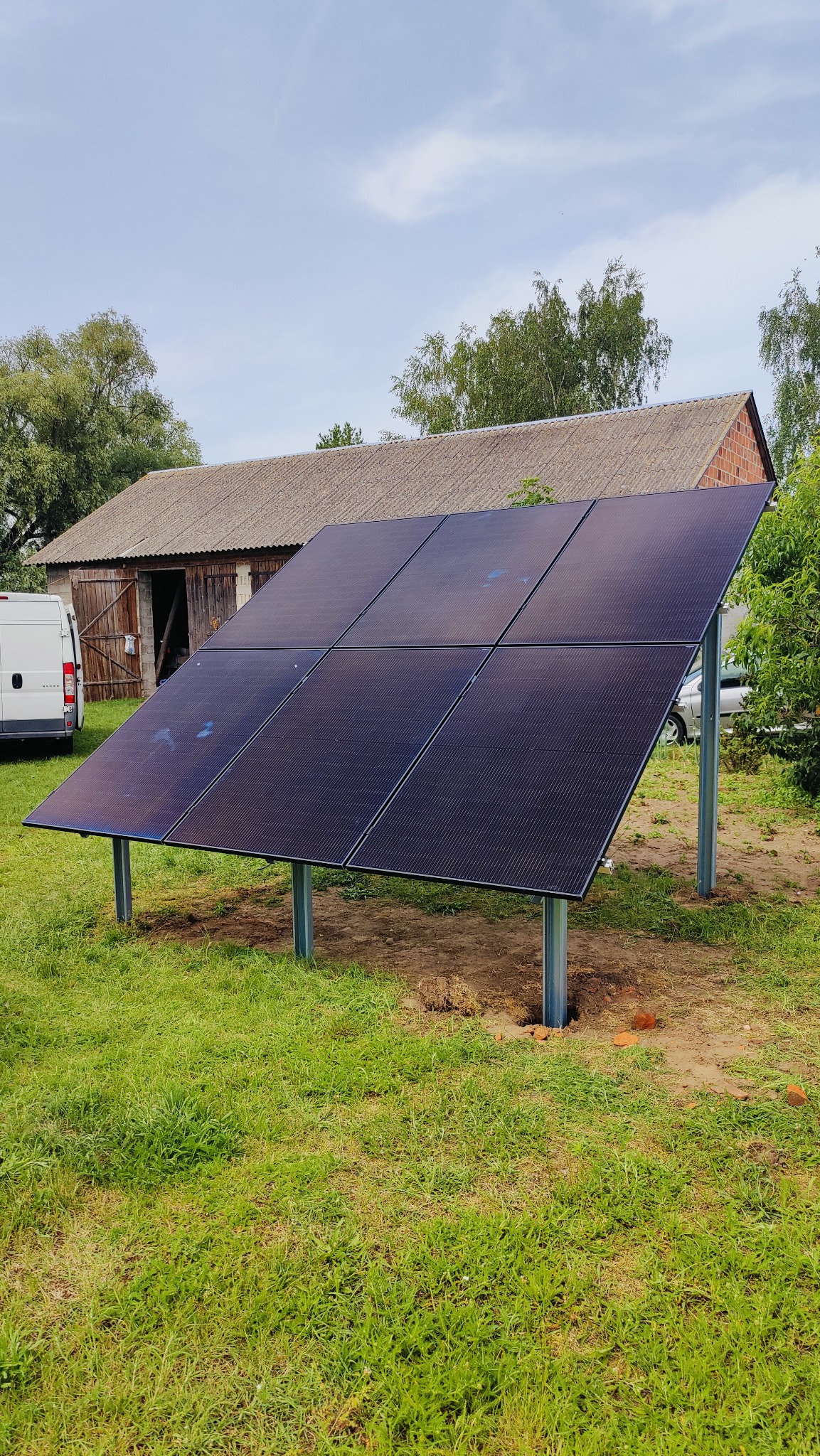Panele słoneczne na metalowej konstrukcji ustawione na trawie, w tle budynek gospodarczy i biały bus.