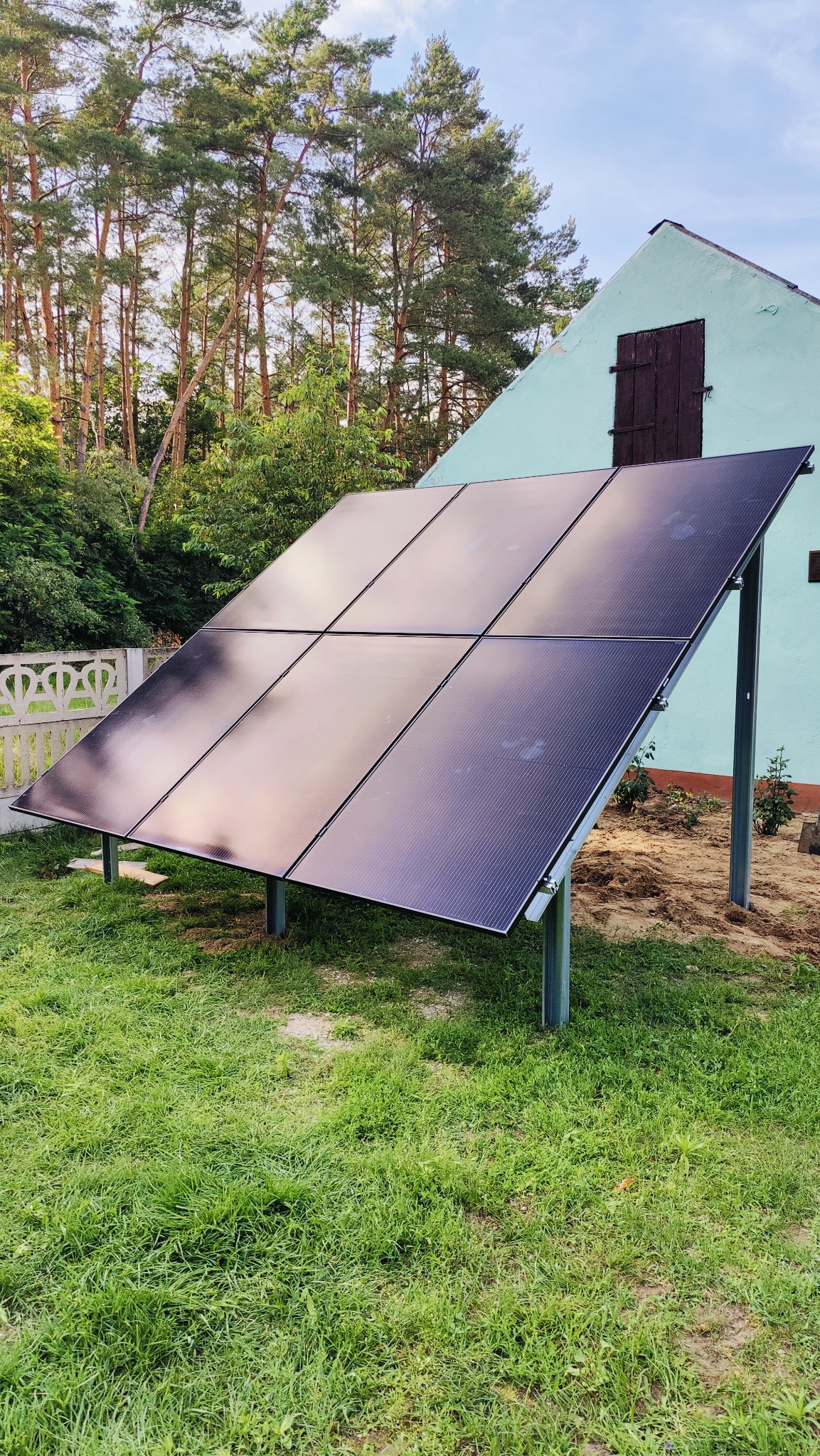 Panele słoneczne zamontowane na trawniku obok turkusowego budynku z drewnianymi okiennicami, w tle wysokie drzewa.