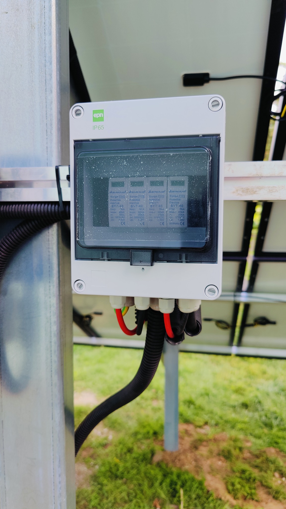 Szczegółowe ujęcie skrzynki zabezpieczeń przepięciowych z oznaczeniem IP65, zamontowanej na metalowym słupku instalacji fotowoltaicznej, widoczne okablowanie i fragment paneli słonecznych w tle.