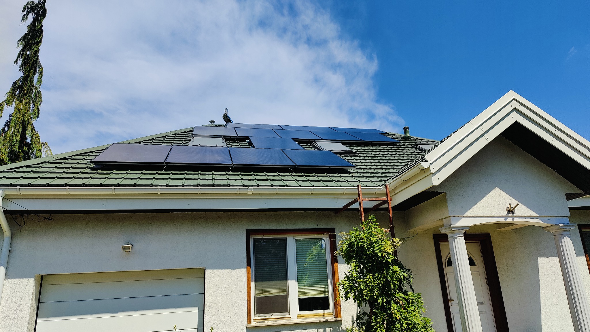 Panele słoneczne zamontowane na zielonym dachu domu jednorodzinnego, widoczne drabiny oparta o ścianę, błękitne niebo z chmurami w tle.