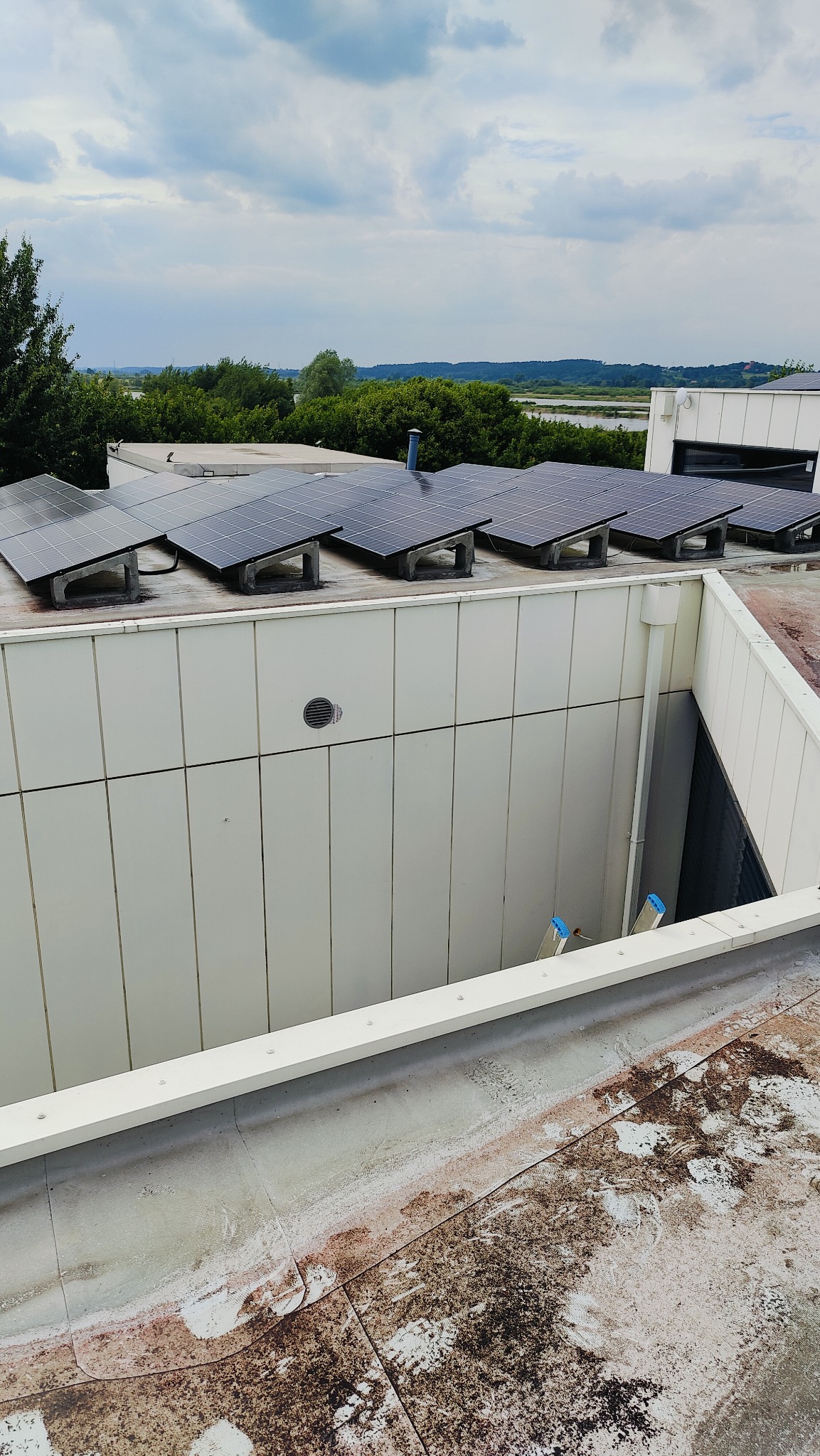 Panele fotowoltaiczne zamontowane na płaskim dachu budynku przemysłowego z widoczną wentylacją i drabiną w zagłębieniu dachu.