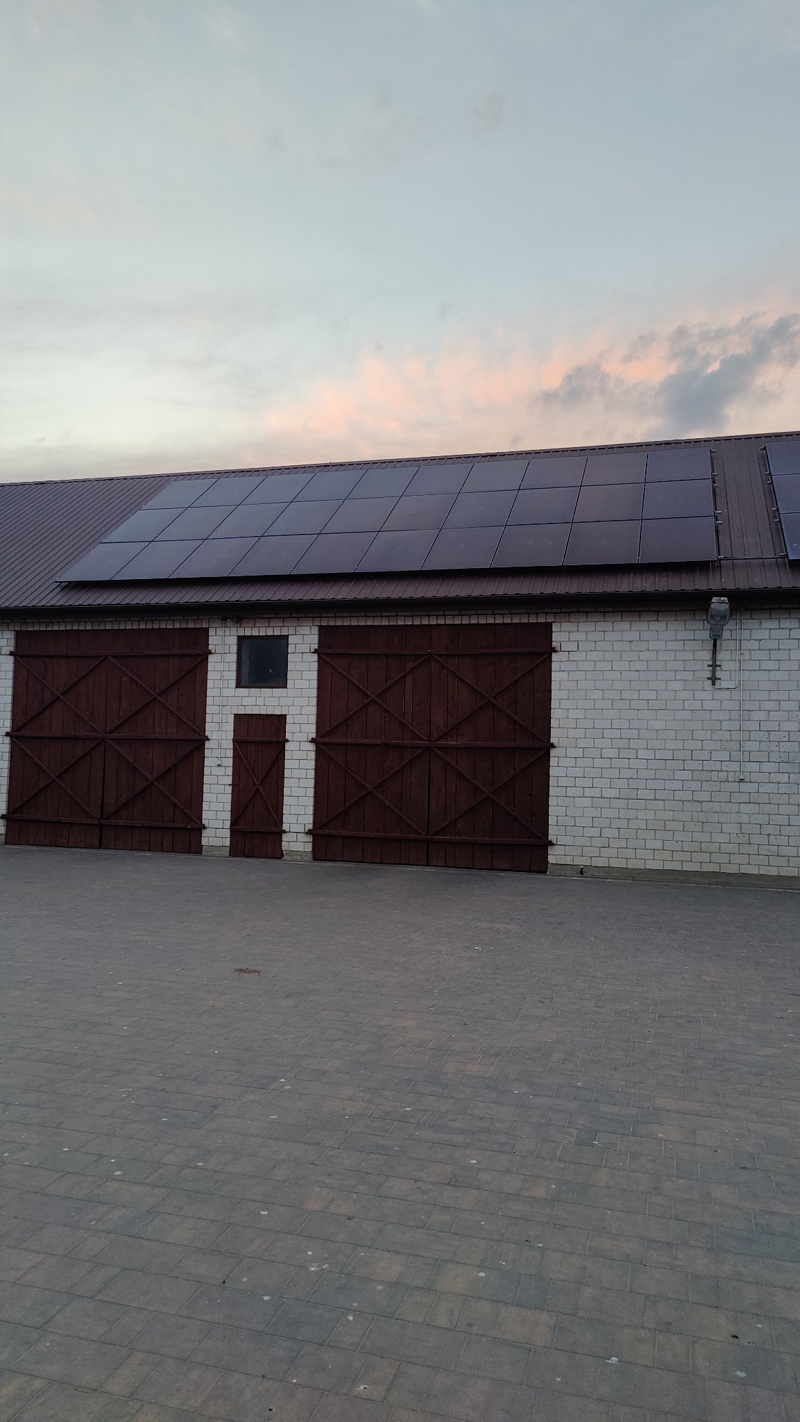Biały budynek gospodarczy z brązowymi, drewnianymi wrotami i panelami słonecznymi na dachu, sfotografowany o zmierzchu.