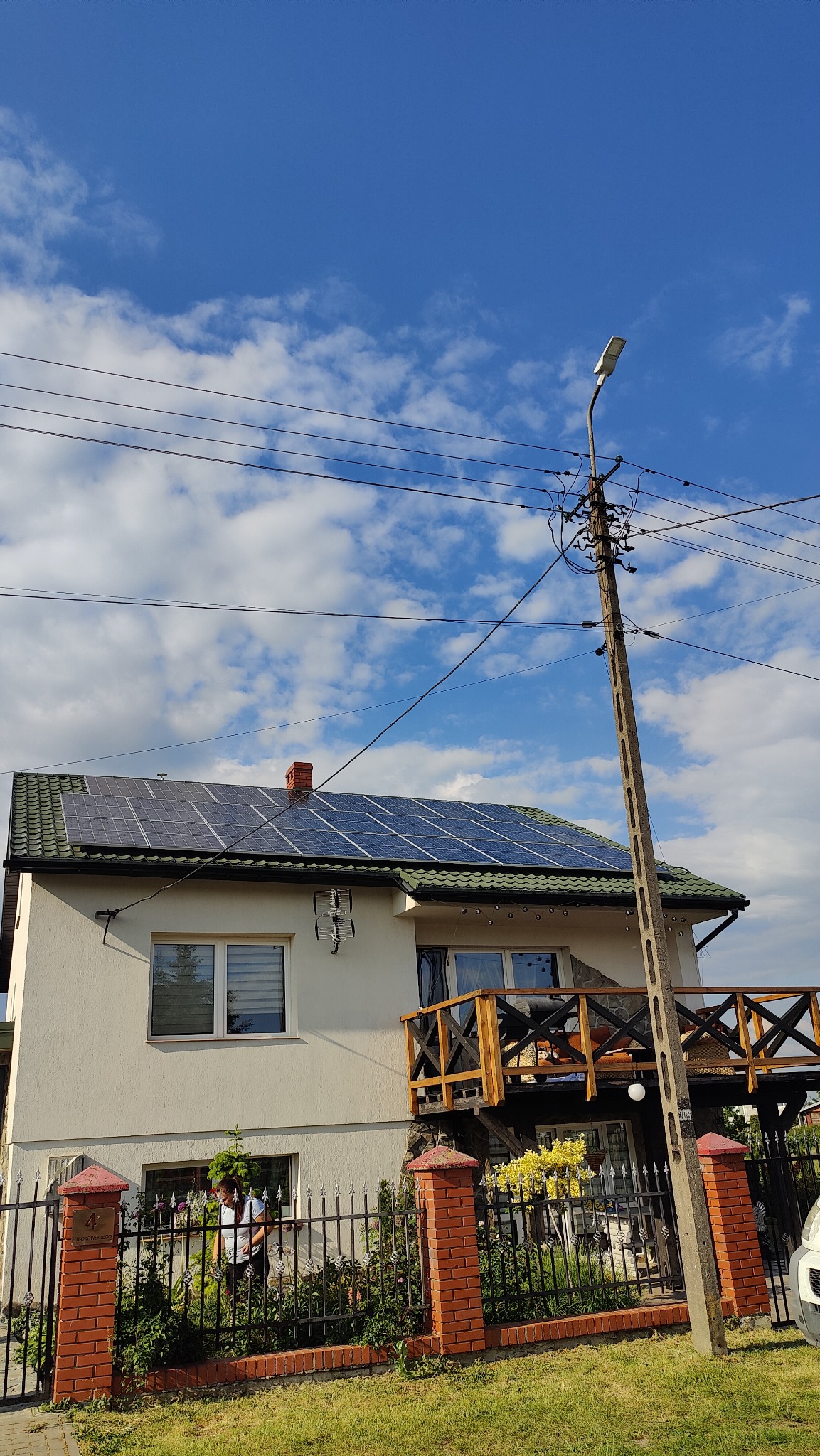 Biały dom z panelem słonecznym na dachu i drewnianym balkonem, widok z dołu na tle błękitnego nieba z chmurami, z przodu ogrodzenie z cegły i żelaza.