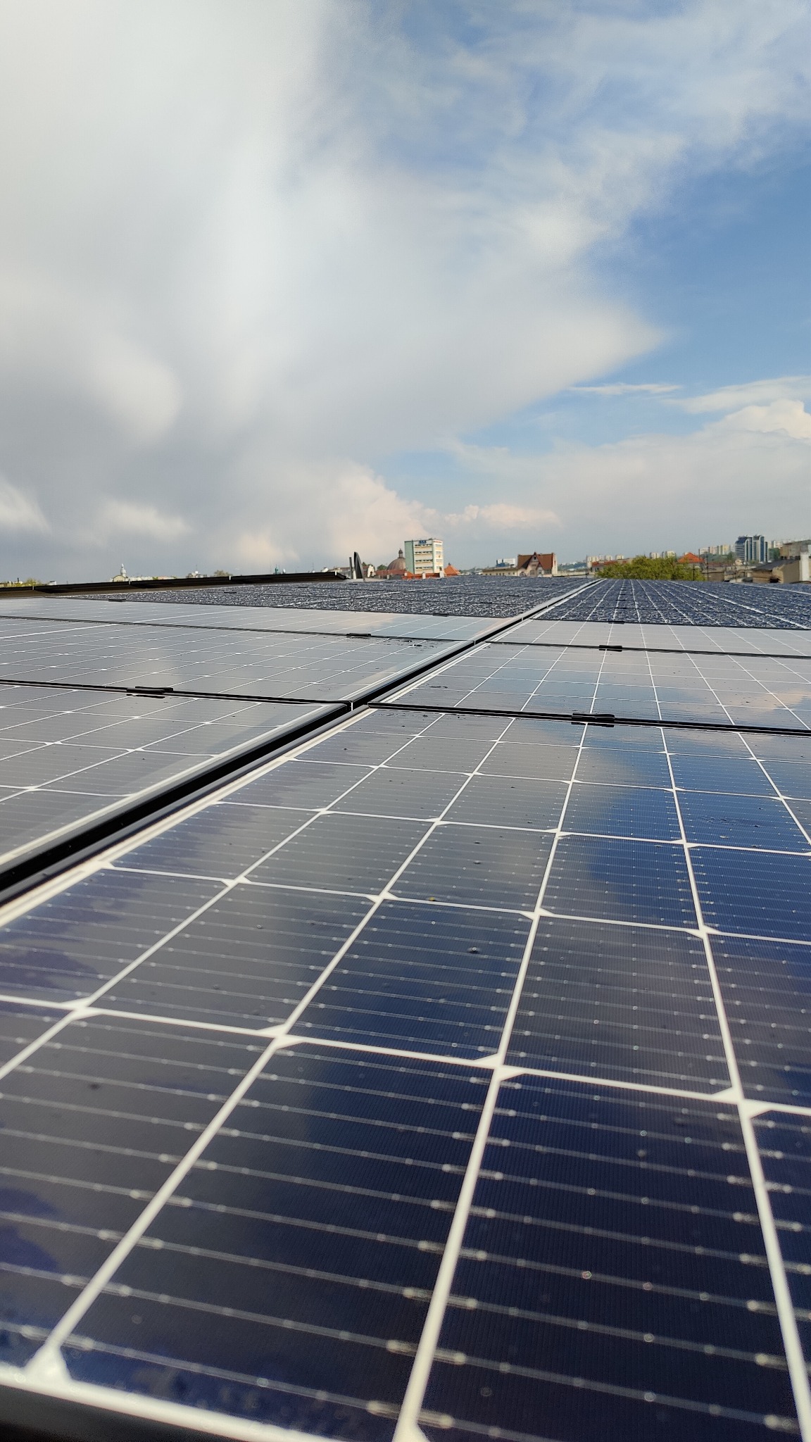 Panel słoneczny na dachu budynku, z widocznym odbiciem chmur i fragmentem panoramy miasta w tle.