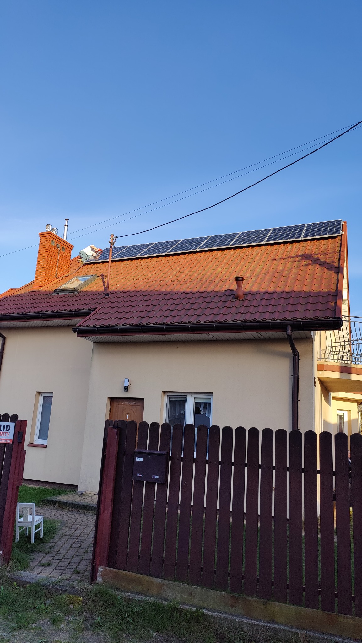 Montaż paneli słonecznych na dachu domu jednorodzinnego z czerwoną dachówką, widoczny pracownik w białej koszulce na dachu, błękitne niebo w tle.