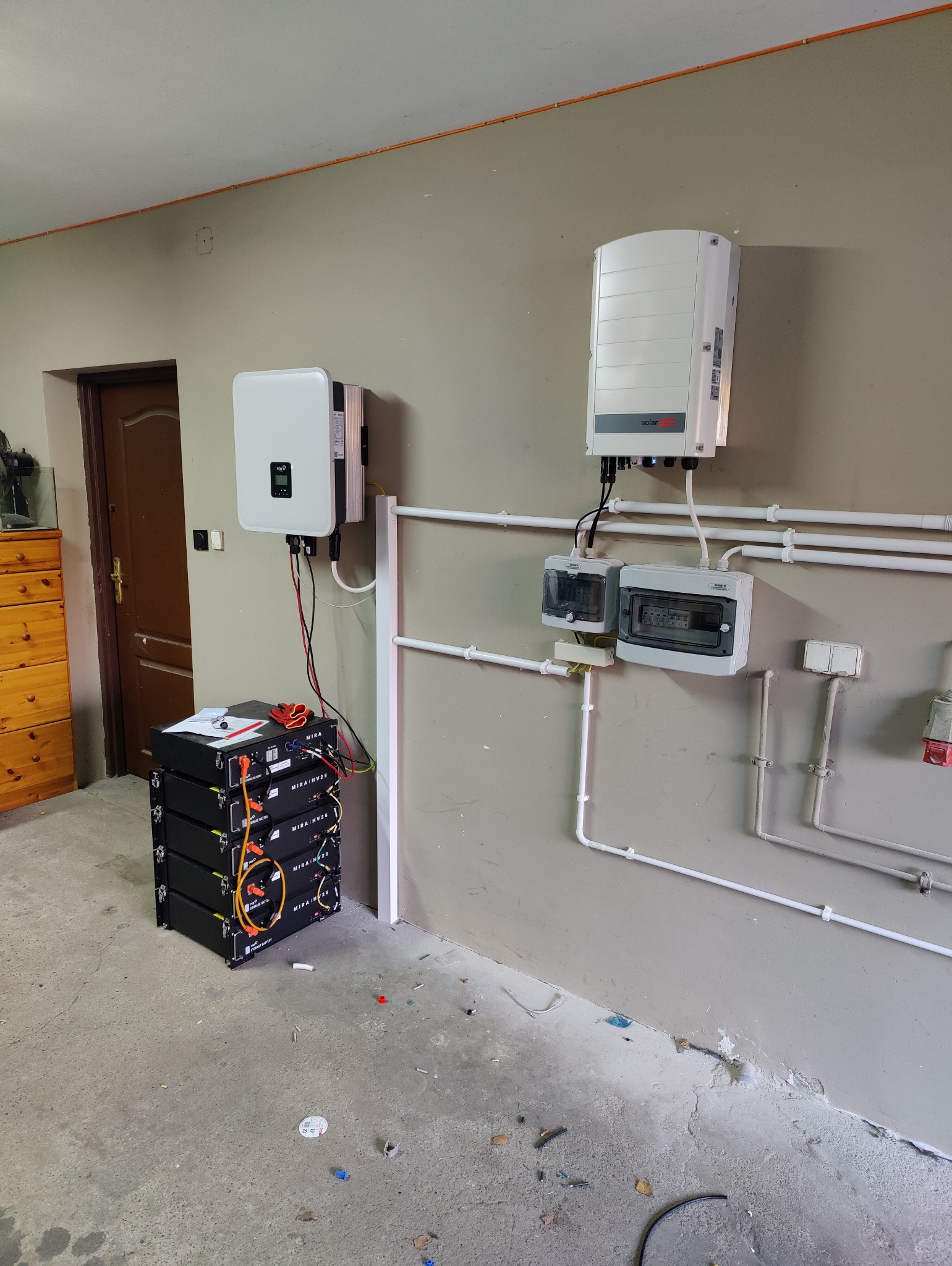 Cztery czarne moduły bateryjne MIRA NV25 ułożone w stos, obok dwa białe inwertery fotowoltaiczne na ścianie, połączone przewodami i białe rury instalacyjne na szarej ścianie w pomieszczeniu.