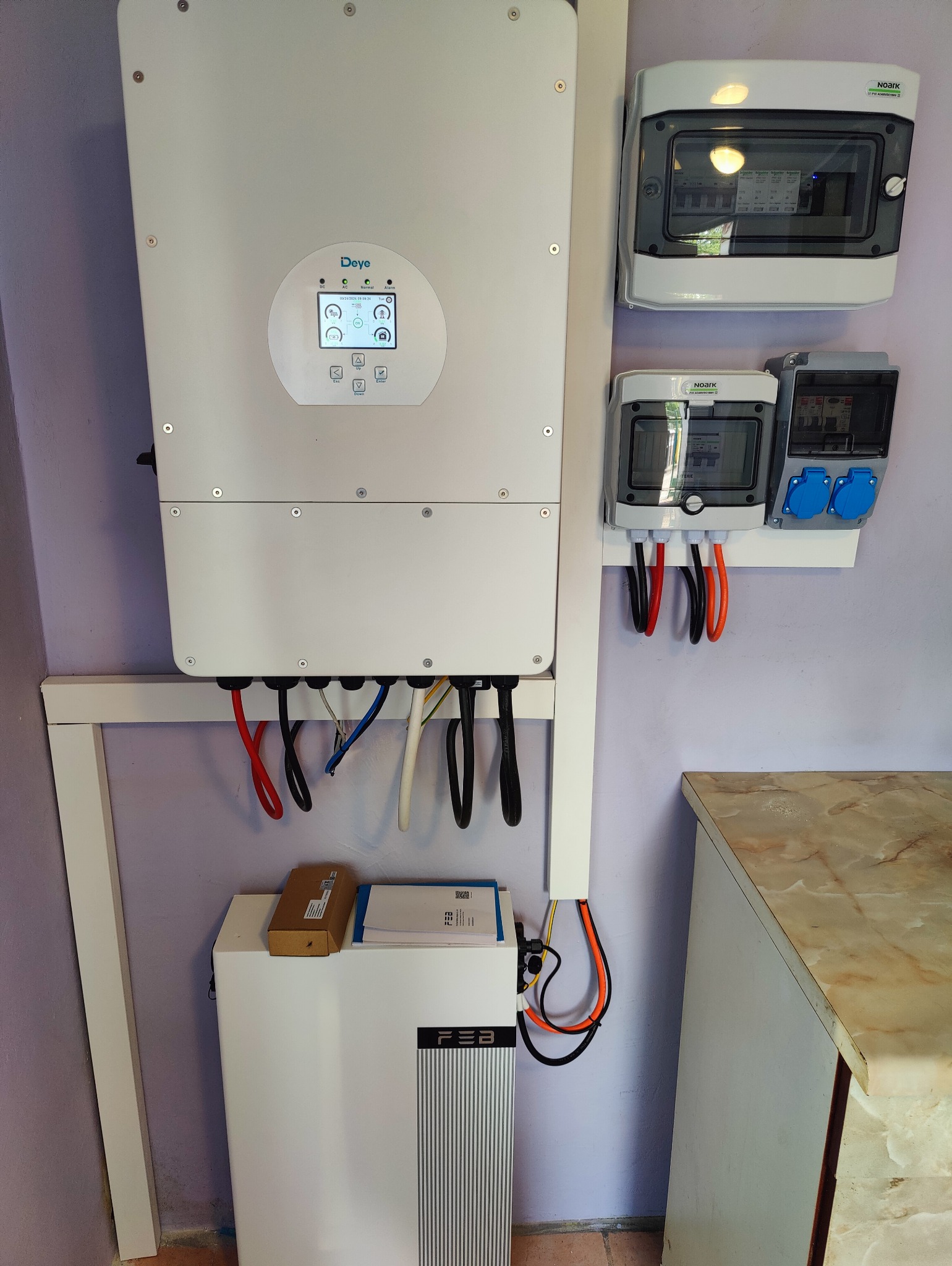 Biały inwerter Deye z wyświetlaczem, zamontowany na ścianie obok skrzynki bezpiecznikowej i gniazd. Pod spodem znajduje się biała bateria FEB z dokumentacją i kartonem na górze. Widoczne przewody...