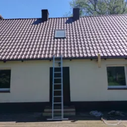 Dom w Oławie z nowym, brązowym dachem z dachówki ceramicznej, widok na elewację z oknami i drabiną opartą o ścianę, przygotowania do prac na wysokości.