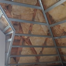 Usługi Remontowe i Wykończeniowe - Skośny sufit poddasza w trakcie montażu: widoczna metalowa konstrukcja, izolacja z wełny mineralnej i fragment ściany z płyty gipsowo-kartonowej przy oknie.