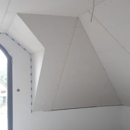 Usługi Remontowe i Wykończeniowe - Montaż płyt kartonowo-gipsowych na skośnym poddaszu, widoczne łączenia płyt i wkręty, surowy betonowy podkład, zabezpieczenie folią wzdłuż ściany, okno dachowe w trakcie obróbki.