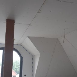 Usługi Remontowe i Wykończeniowe - Poddasze w trakcie wykańczania płytami kartonowo-gipsowymi, widoczny drewniany słup, okno dachowe i okno w szczycie, szpachlowane łączenia płyt.