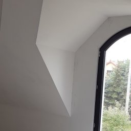 Usługi Remontowe i Wykończeniowe - Wnętrze poddasza po pracach z płyt kartonowo-gipsowych, widoczne skośne ściany, sufit z wystającym kablem i okno z czarną ramą, na podłodze butelka wody.