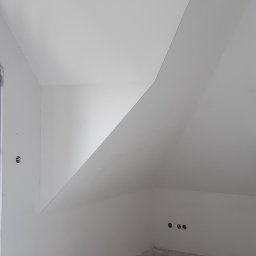 Usługi Remontowe i Wykończeniowe - Poddasze w stanie deweloperskim, białe ściany i sufit, widoczne gniazdka elektryczne i wystający przewód z sufitu, surowa wylewka na podłodze.