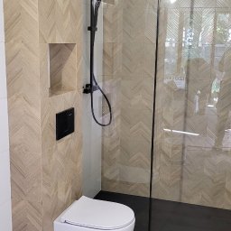 Usługi Remontowe i Wykończeniowe - Nowoczesna łazienka z płytkami w jodełkę na ścianach i podłodze, czarna armatura prysznicowa i podwieszana toaleta z przyciskiem spłukiwania w kolorze czarnym.