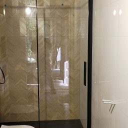 Usługi Remontowe i Wykończeniowe - Nowoczesna łazienka z kabiną prysznicową typu walk-in z czarną ramą, toaleta podwieszana i płytki imitujące drewno na podłodze i ścianie prysznica ułożone we wzór jodełki.