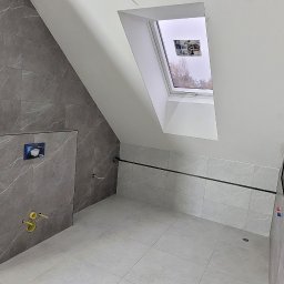 Usługi Remontowe i Wykończeniowe - Wykończona łazienka na poddaszu w szarościach z marmurkowym wzorem płytek, widoczne przyłącza wodne i okno dachowe, minimalistyczny styl.