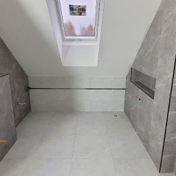 Usługi Remontowe i Wykończeniowe - Szara łazienka z marmurowymi płytkami, oknem dachowym i wnęką prysznicową, widoczne przyłącza instalacyjne.