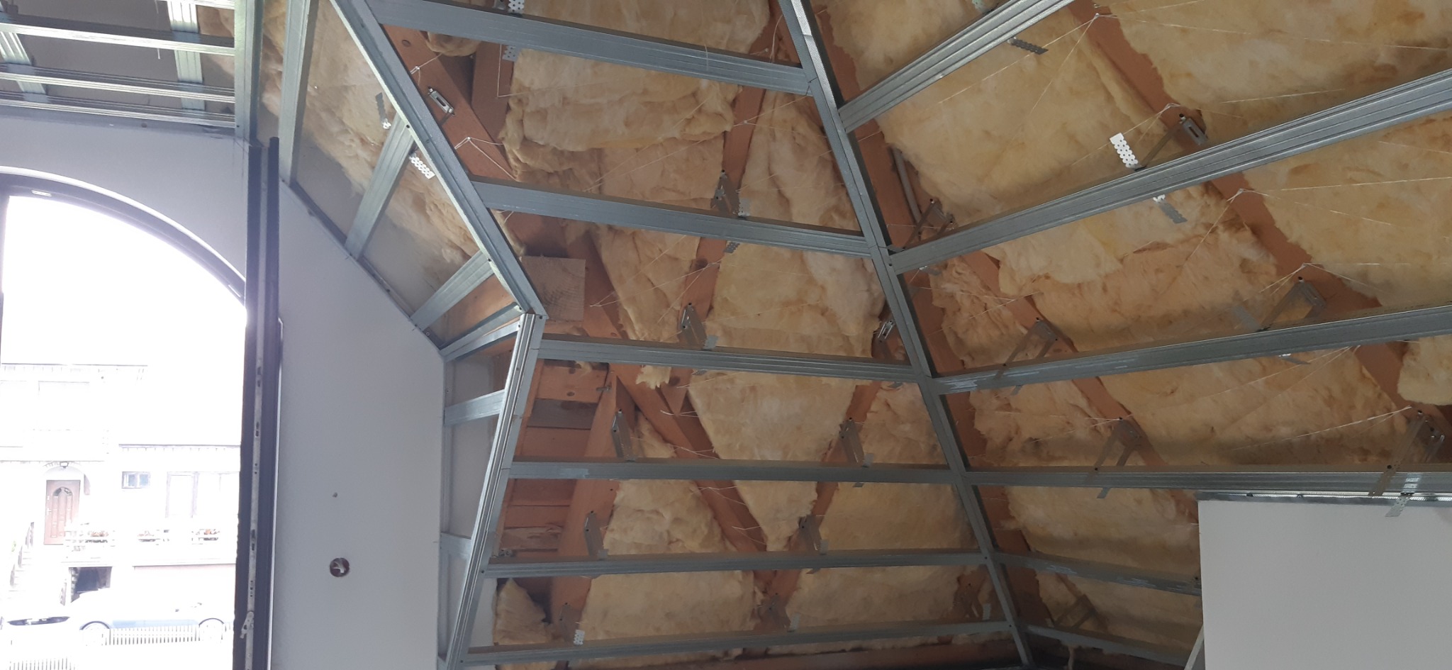 Skośny sufit poddasza w trakcie montażu: widoczna metalowa konstrukcja, izolacja z wełny mineralnej i fragment ściany z płyty gipsowo-kartonowej przy oknie.