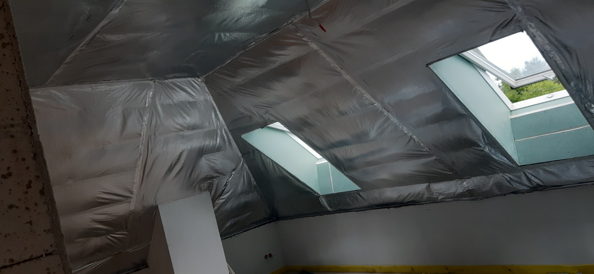 Poddasze w trakcie prac wykończeniowych: folia termoizolacyjna na skośnym dachu, okna dachowe z widokiem na zieleń, fragment ściany z widocznymi śladami wilgoci.