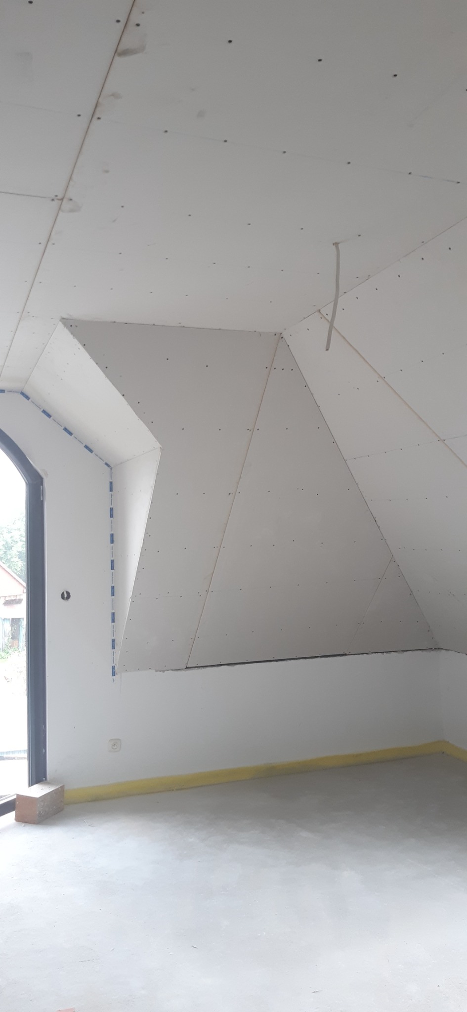 Montaż płyt kartonowo-gipsowych na skośnym poddaszu, widoczne łączenia płyt i wkręty, surowy betonowy podkład, zabezpieczenie folią wzdłuż ściany, okno dachowe w trakcie obróbki.