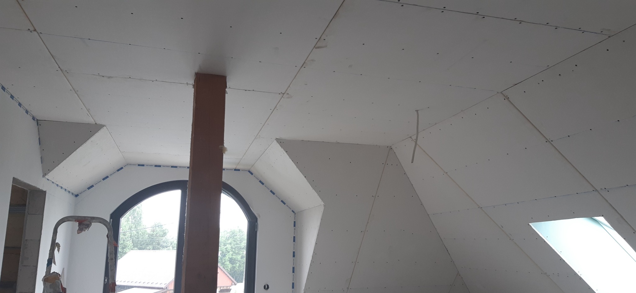 Poddasze w trakcie wykańczania płytami kartonowo-gipsowymi, widoczny drewniany słup, okno dachowe i okno w szczycie, szpachlowane łączenia płyt.