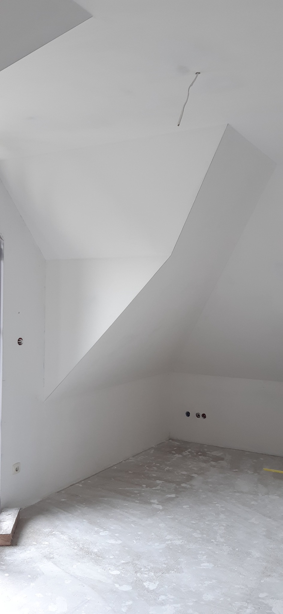 Poddasze w stanie deweloperskim, białe ściany i sufit, widoczne gniazdka elektryczne i wystający przewód z sufitu, surowa wylewka na podłodze.