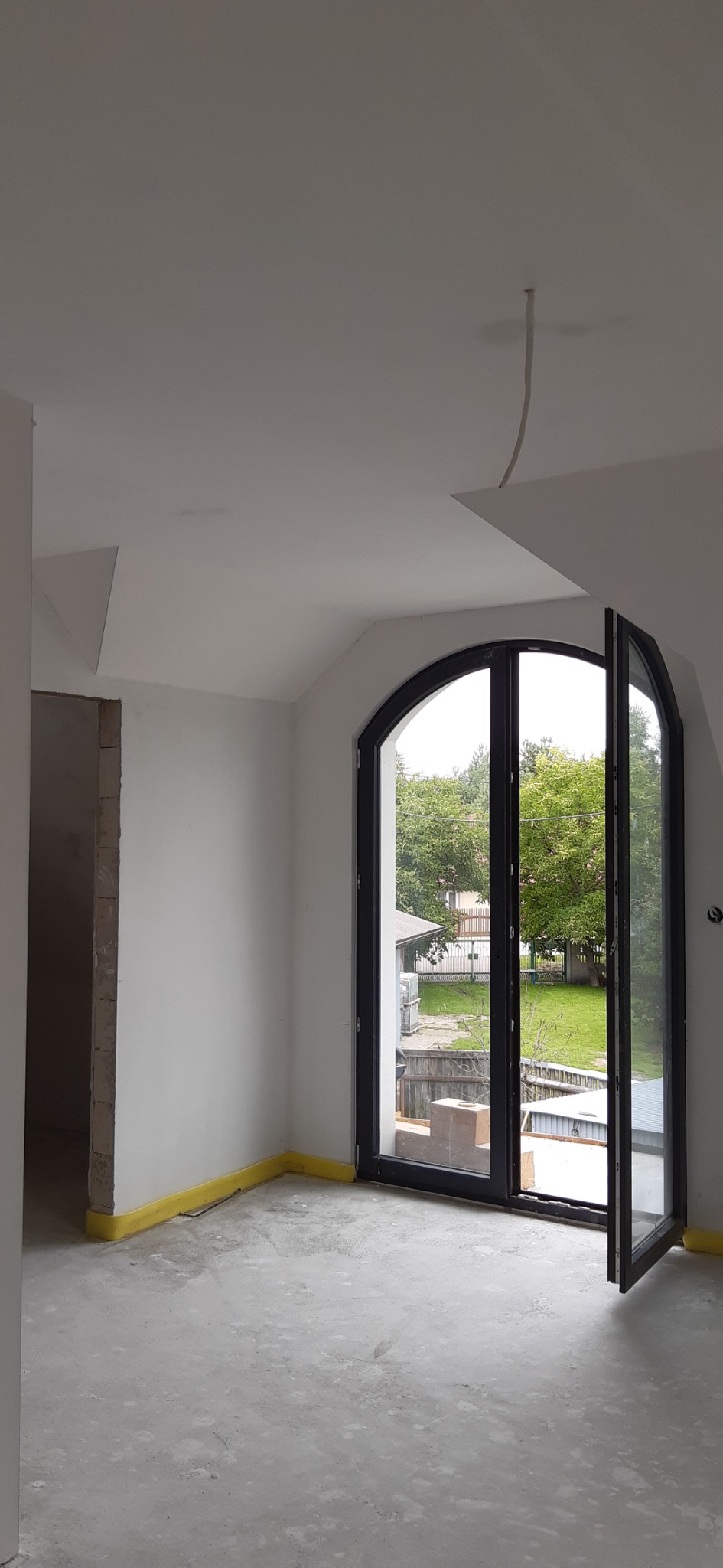 Wnętrze pokoju w trakcie remontu z dużym, łukowym oknem balkonowym, zabezpieczona żółtą folią podłoga, surowe ściany i wystający kabel z sufitu.