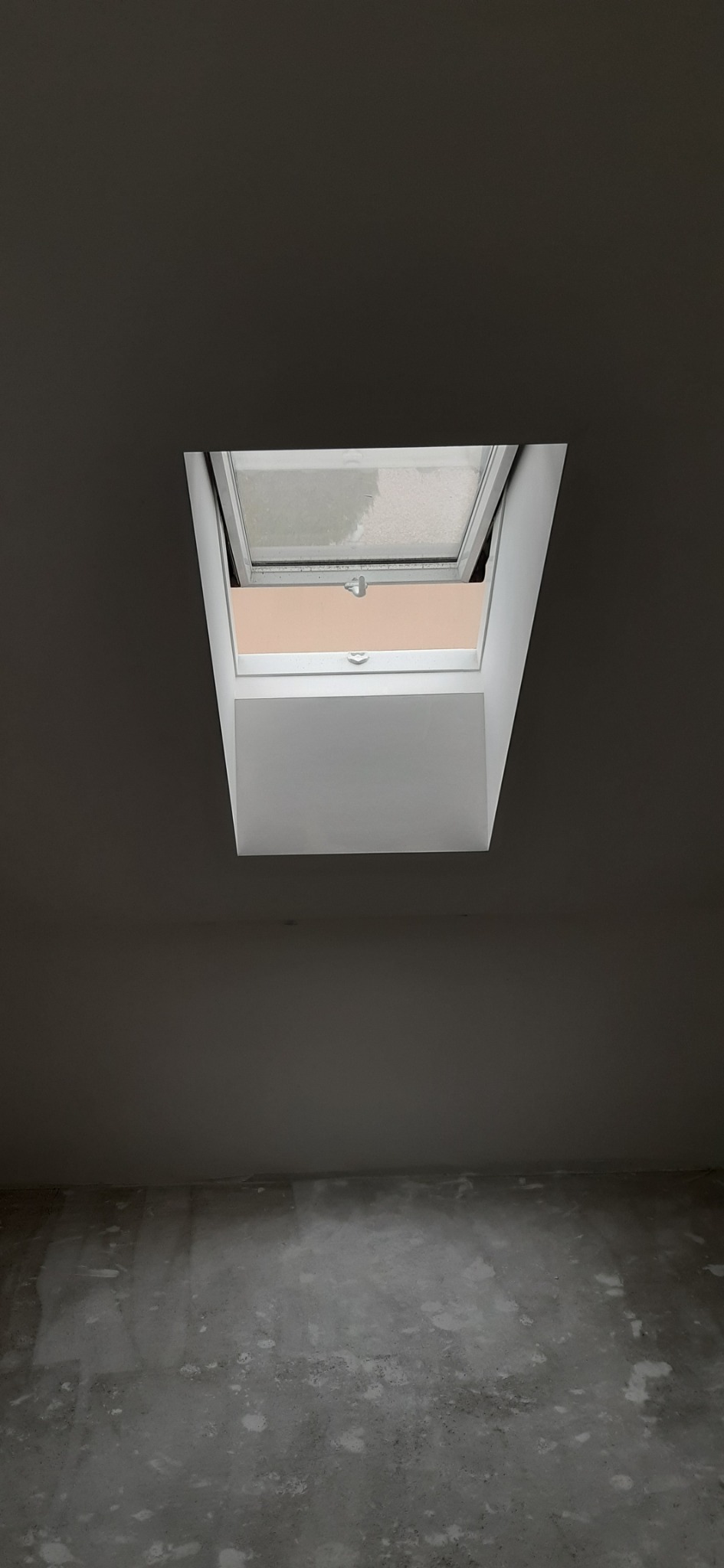 Otwarty okno dachowe w jasnej ramie, widoczne na tle ciemnoszarego sufitu i ściany, surowa podłoga w trakcie prac wykończeniowych.