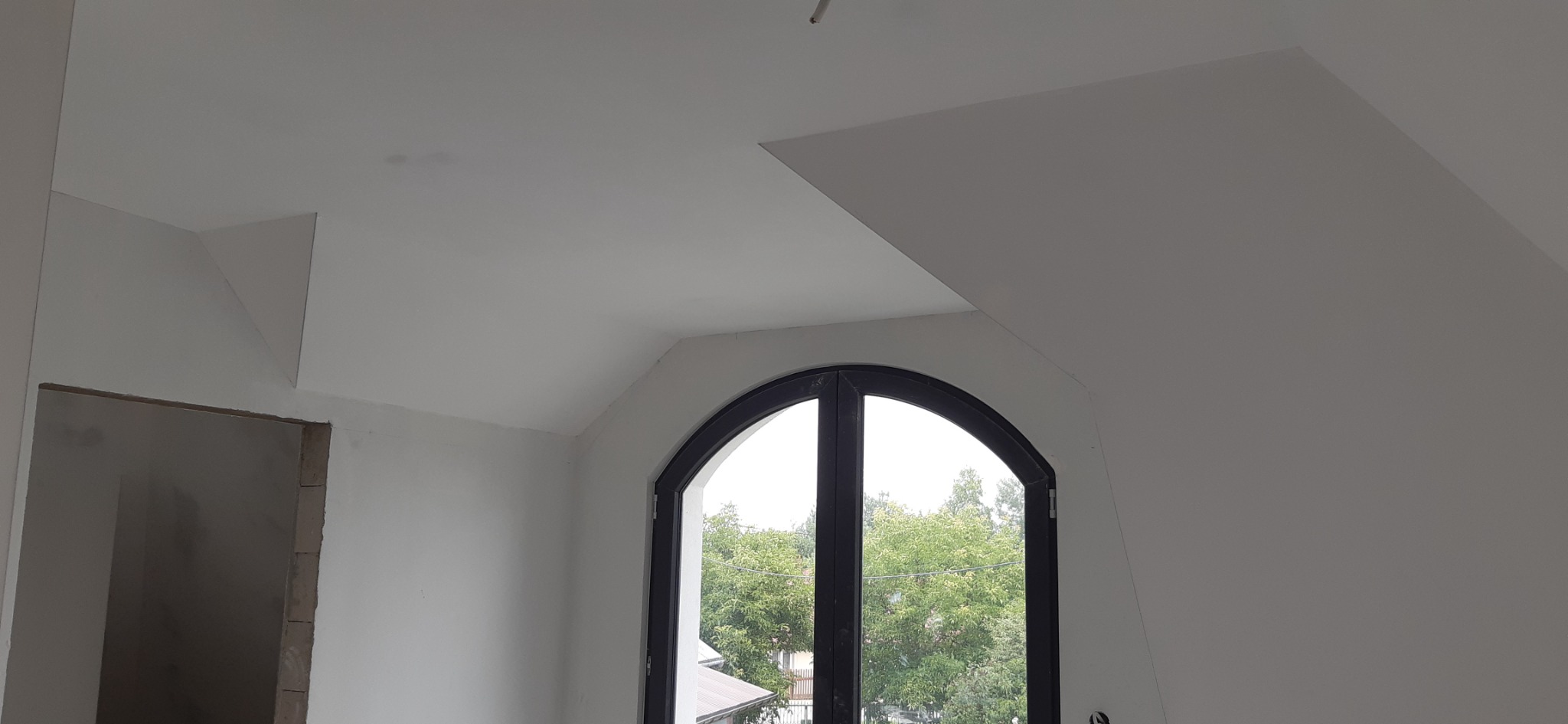 Wnętrze poddasza w trakcie prac, widoczne fragmenty ścian i sufitu po szpachlowaniu, okno łukowe z czarną ramą, surowy otwór drzwiowy.