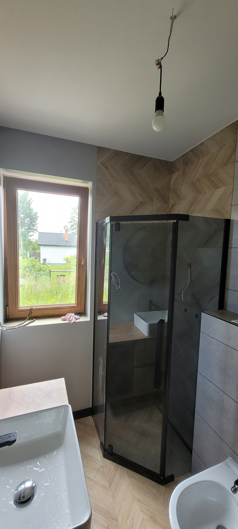 Nowoczesna łazienka z kabiną prysznicową typu walk-in, umywalką nablatową, bidetem i płytkami imitującymi drewno na ścianie, z niedokończoną instalacją oświetleniową.