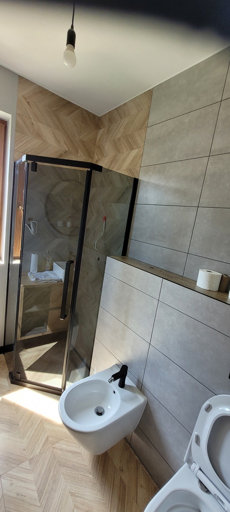 Nowoczesna łazienka po remoncie: kabina prysznicowa z czarnymi ramami, bidet z czarną armaturą, toaleta, płytki imitujące drewno na podłodze i ścianie, szare płytki ścienne.
