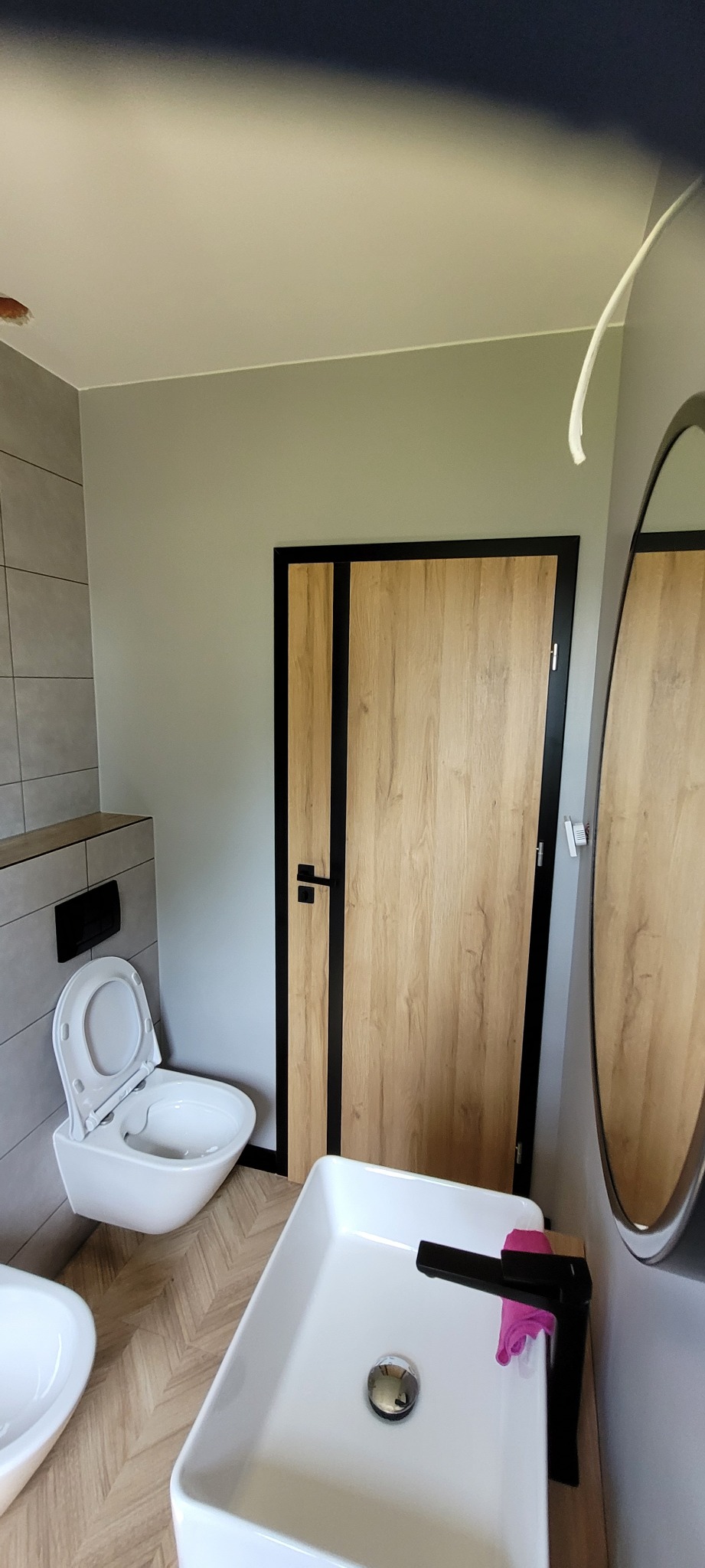 Nowoczesna łazienka z podwieszaną toaletą, prostokątną umywalką z czarną armaturą i drewnianymi drzwiami z czarnymi wstawkami, widoczne odbicie umywalki w lustrze, na umywalce różowa ściereczka.