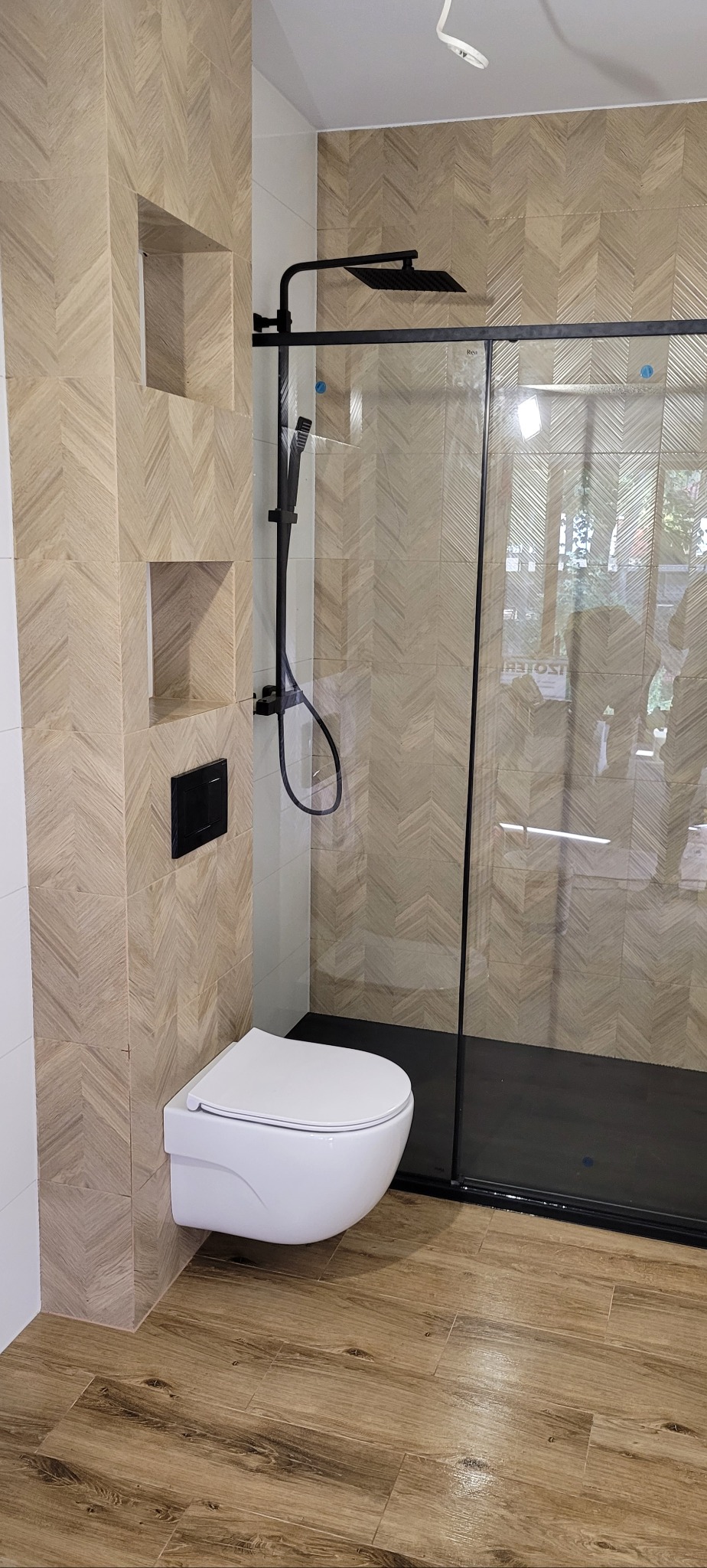 Nowoczesna łazienka z płytkami w jodełkę na ścianach i podłodze, czarna armatura prysznicowa i podwieszana toaleta z przyciskiem spłukiwania w kolorze czarnym.