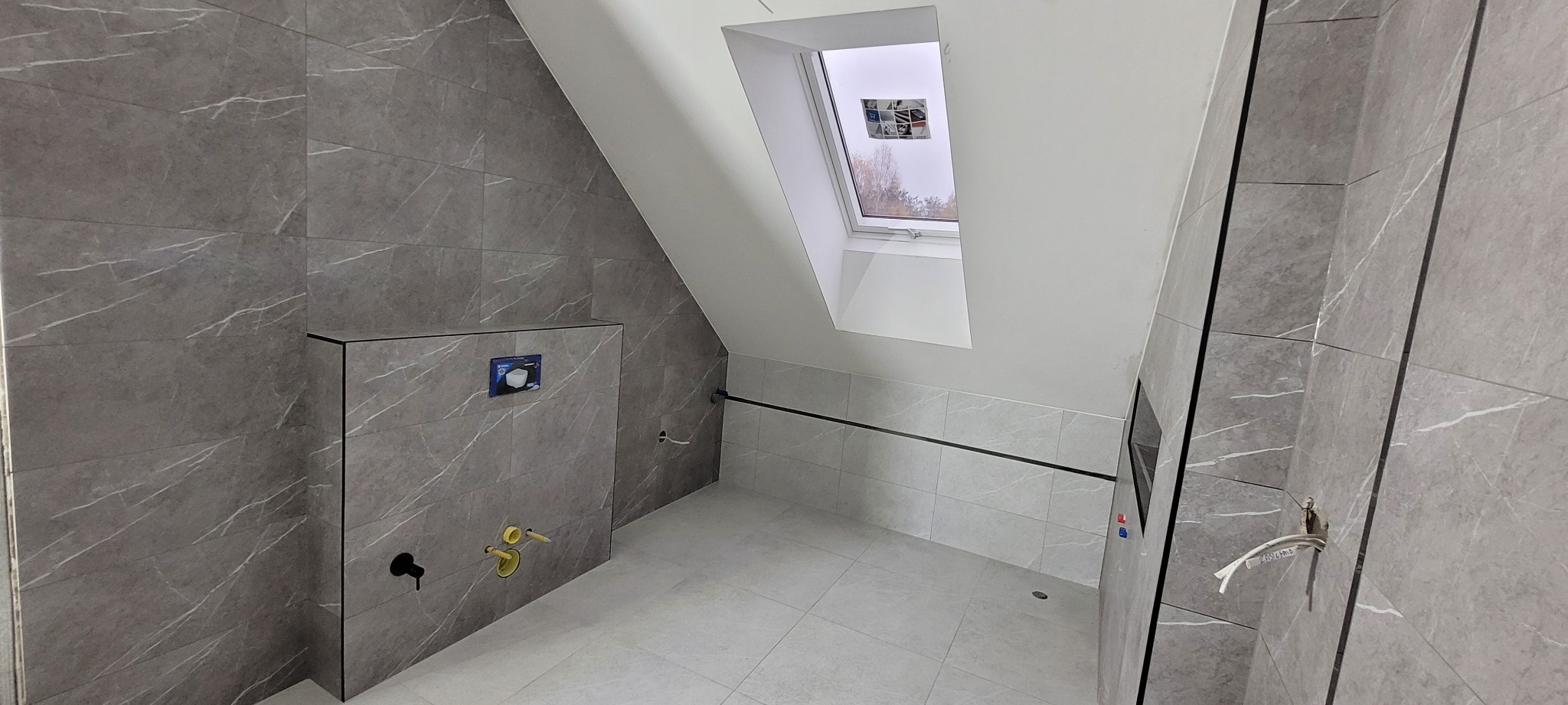 Wykończona łazienka na poddaszu w szarościach z marmurkowym wzorem płytek, widoczne przyłącza wodne i okno dachowe, minimalistyczny styl.