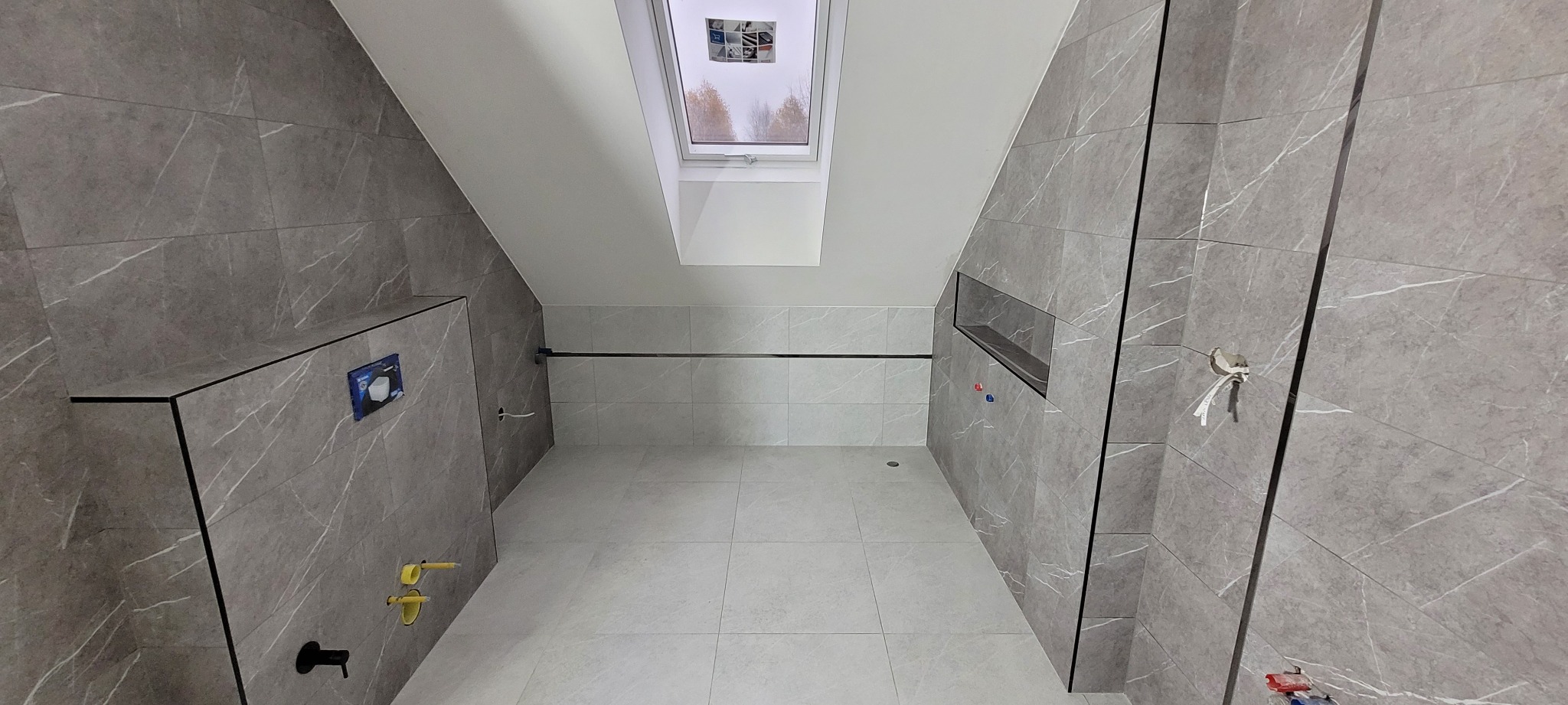 Szara łazienka z marmurowymi płytkami, oknem dachowym i wnęką prysznicową, widoczne przyłącza instalacyjne.