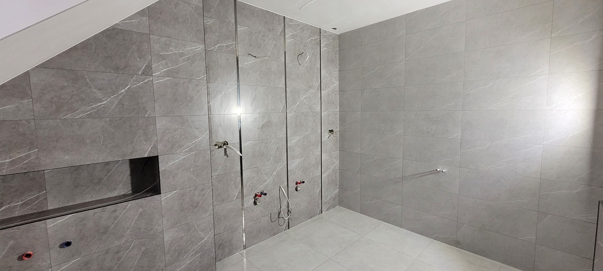 Ściany i podłoga wyłożone szarymi płytkami imitującymi marmur, widoczne instalacje wodne w trakcie montażu, wnęka wykończona płytkami na ścianie po lewej stronie.